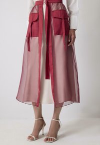 Femme portant une jupe bordeaux transparente avec de grandes poches avant, une ceinture nouée, une robe blanche en dessous, et des sandales à talons blancs, se tenant sur un sol blanc.
