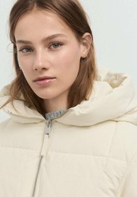 Chaqueta acolchada en beige claro con capucha, que presenta un cierre frontal y una textura acolchada. Debajo, se puede ver un suéter de punto gris.