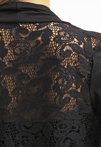 Svart topp med intrikata spetsdetaljer på baksidan, med blommönster och en slät tygfront. Texturerad, halvgenomskinlig spets kontrasterar med det solida materialet.