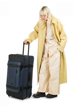 Grande valigia trolley blu navy con accenti neri, tessuto resistente, molteplici scomparti e manico telescopico, accanto a una figura con cappotto giallo.