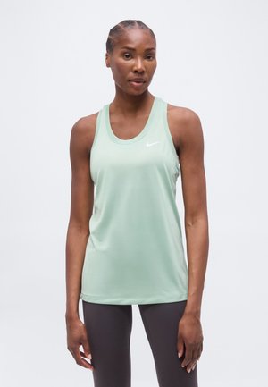 Femme aux cheveux tressés portant un débardeur de sport Nike vert clair sans manches et un legging foncé, debout devant un fond clair uni.