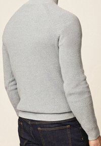 Uomo che indossa un maglione turtleneck a maglia grigio chiaro e jeans in denim blu scuro, visto di spalle e dalla vita in su.