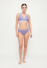 Woman standing barefoot against white background wearing sheer blue lace bra and matching underwear, looking forward with neutral expression.