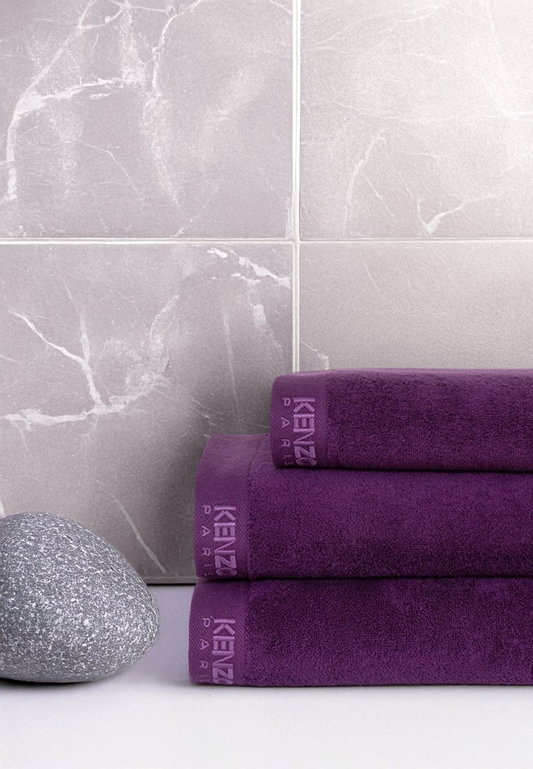 Tres toallas apiladas de color púrpura, hechas de un tejido suave, con una superficie texturizada y la marca KENZO. En primer plano, una piedra gris suave. El fondo cuenta con baldosas de mármol gris claro.