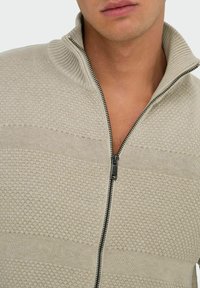 Pull zippé beige avec un col côtelé, motif texturé et rayures horizontales contrastantes. Présente un détail de fermeture éclair foncé.