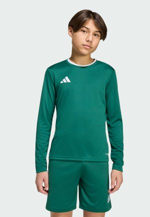 Young person standing, wearing a green long-sleeve sports shirt and matching shorts with white Adidas logos on each.