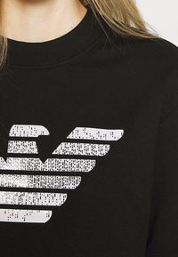 Svart sweatshirt med rundad halsringning som har en stor, strukturerad vit logotyp som liknar stiliserade vingar på framsidan.