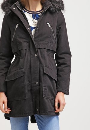 Femme portant un parka noir avec une capuche bordée de fourrure, des poches zippées sur la poitrine, des poches sur les hanches avec rabat et un jean bleu.