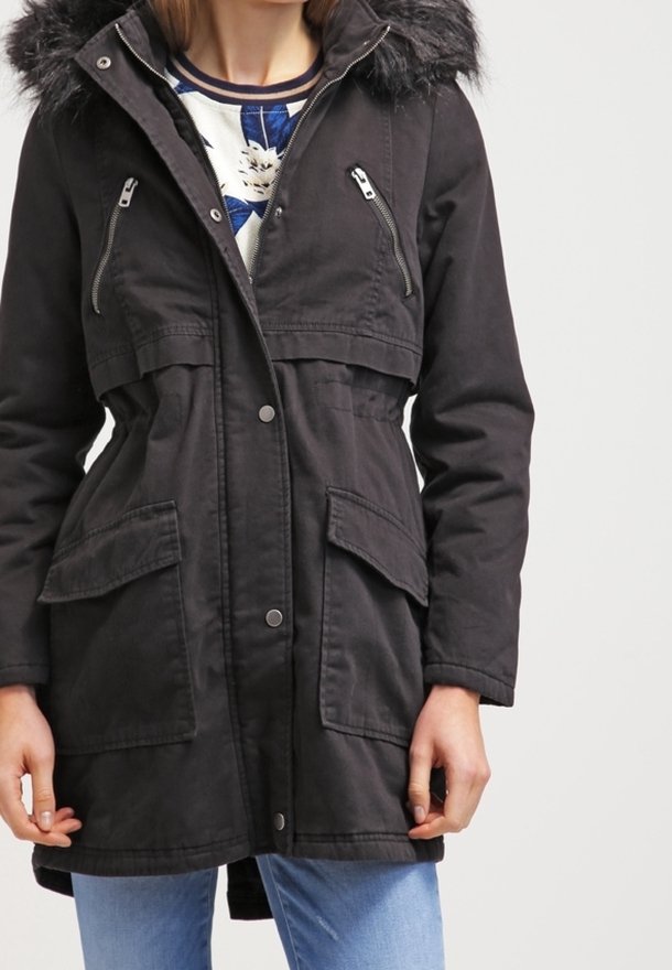 Kobieta ubrana w czarną parkę z kapturem wykończonym futrem, zamkniętymi kieszeniami na piersi, kieszeniami na biodrach z klapami i niebieskimi dżinsami.