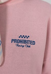 Vêtement en tricot rose clair avec texte brodé en bleu « PROHIBITED Racing Club » et accents à motif à damier sur l'encolure.
