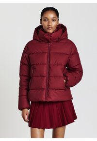 Donna che indossa un giubbotto imbottito bordeaux con un colletto alto e cappuccio, abbinato a una gonna plissettata coordinata, in piedi di fronte a uno sfondo bianco.