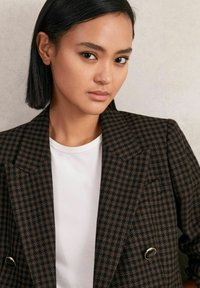 Blazer à carreaux marron avec des revers surdimensionnés, conception croisée, et boutons contrastants, associé à un t-shirt blanc à col rond.