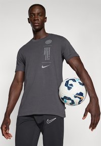 Šedé tričko s krátkym rukávom s potlačou a logom Nike, doplnené čiernymi športovými nohavicami a bielou a modrou futbalovou loptou.