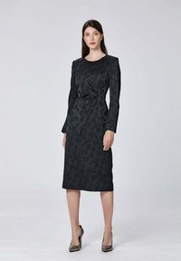 Robe noire jusqu'aux genoux avec des manches longues et un motif floral, dotée d'une taille cintrée et d'un col rond. Chaussures à talons hauts.