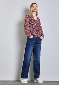 Langarm, doorzichtig, bordeauxrood bovenstuk met abstract patroon, gecombineerd met hooggesneden, donkerblauwe rechte jeans en witte sneakers.