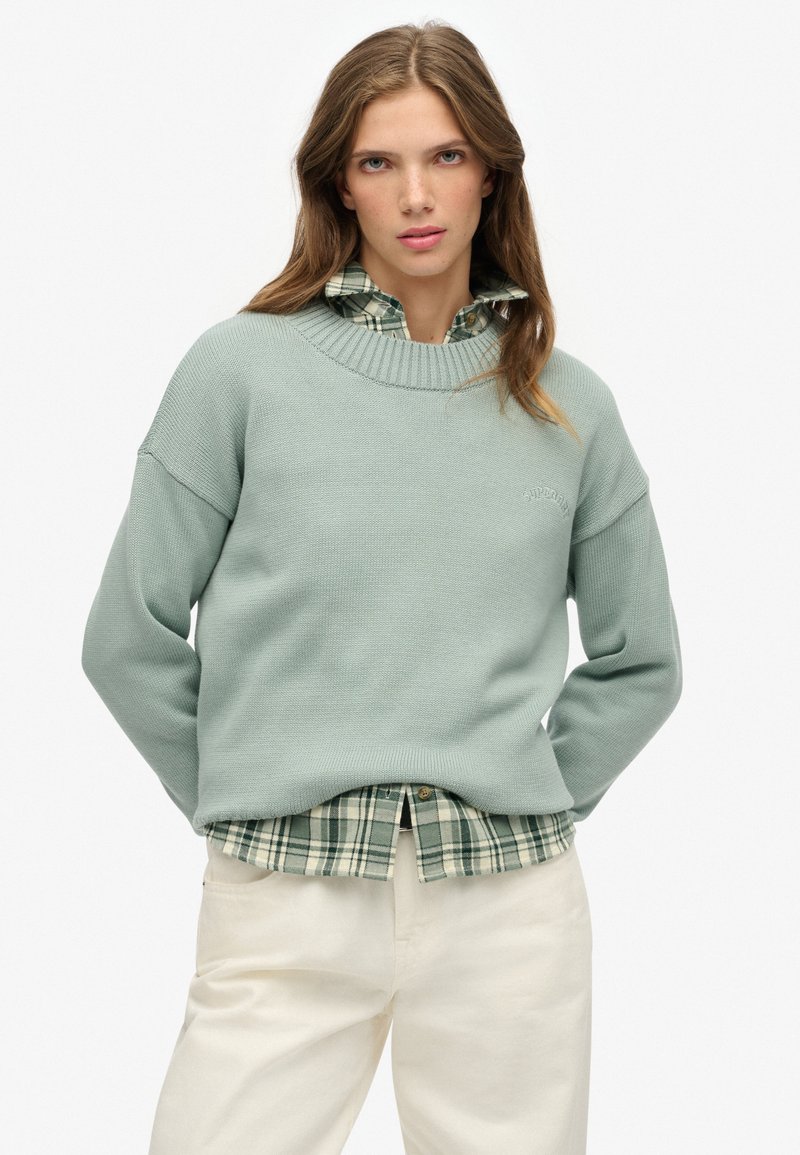 Joven mujer vestida con un suéter verde claro sobre una camisa de cuadros verdes y pantalones color crema, de pie con las manos detrás de la espalda.