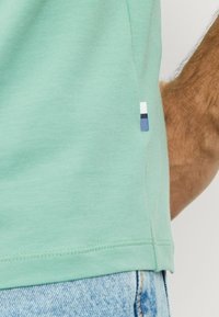 Mint green cotton t-shirt with short sleeves, featuring a stitched hem and a small, multi-colored logo tag at the side seam.