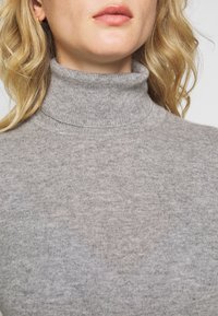 Lichtgrijze coltrui gemaakt van een zachte, gebreide stof met textuur. Heeft een aansluitend ontwerp en een geribbelde kraag. Close-up weergave.