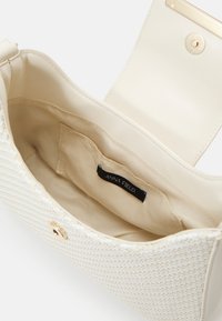 Biała teksturowana torebka z gładką skórzaną klapką, złotymi okuciami i beżowym wnętrzem z etykietą marki. Kompaktowy i strukturalny design.