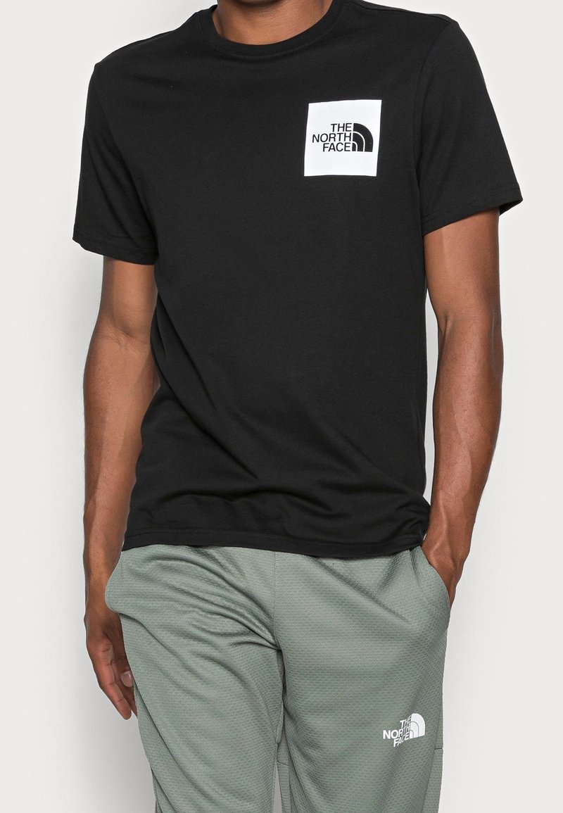 Czarny bawełniany t-shirt z krótkimi rękawami, z białym kwadratowym logo na lewej piersi. Noszony z jasnozielonymi spodniami o fakturowanej strukturze.