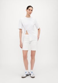 Hvit t-skjorte med kort erme, tvistdetalj og sirkulært utskjæring i midjen, i kombinasjon med hvite shorts og svart-hvite joggesko.
