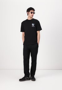 Zwarte katoenen t-shirt met een grafische print. Gepaarde met zwarte broeken en zwarte sneakers. Grijze zonnebril maakt de outfit compleet.