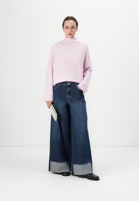 Pulover turtleneck cu model în dungi de culoare roz deschis, asortat cu jeansi din denim întunecat cu picior larg și duble manșete, ținând o poșetă texturată de argint.