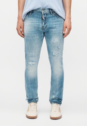 Άνδρας που φοράει ανοιχτό μπλε φθαρμένα τζιν και λευκά αθλητικά παπούτσια, στέκεται μπροστά σε ένα απλό λευκό φόντο με χαλαρά τα χέρια στα πλευρά.