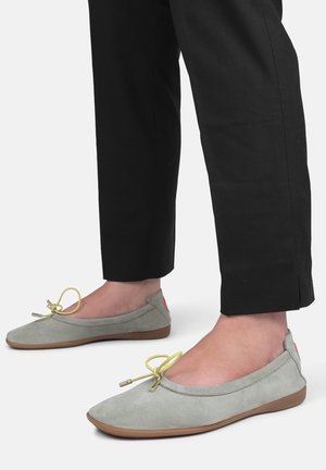 Ballerines en suède gris clair avec un bout rond, des lacets jaunes et une semelle en caoutchouc, associées à un pantalon noir ajusté.