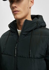 Junger Mann trägt eine schwarze gepolsterte Jacke mit Kapuze und Reißverschluss auf der Vorderseite, welche die Details des Stoffes und der Nähte der Jacke zeigt.