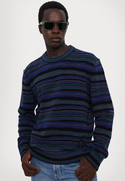 Uomo che indossa occhiali da sole neri, un maglione a righe blu e verdi e jeans azzurri chiari, in piedi con le mani nelle tasche su uno sfondo semplice.