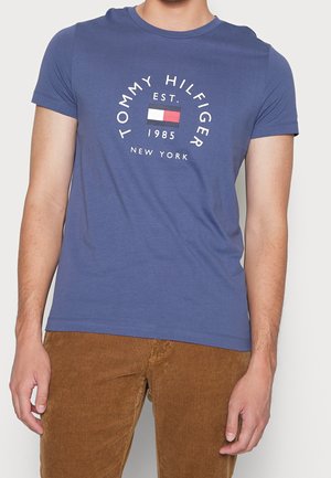 Mężczyzna w niebieskim T-shircie Tommy Hilfiger z logo i napisem „Est. 1985 New York”, w połączeniu z brązowymi sztruksowymi spodniami, stoi na neutralnym tle.