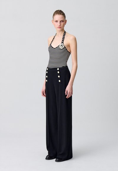 Pantaloni neri a gamba larga con grandi bottoni dorati, abbinati a un top halter a righe bianche e nere con un emblema circolare.
