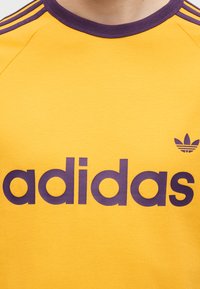 Κίτρινο πουκάμισο Adidas με μοβ κείμενο λογότυπου και έμβλημα trefoil κοντά στο στήθος, με μοβ γιακά και ριγέ λεπτομέρειες στους ώμους.