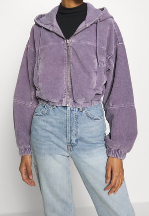 Person trägt eine bauchfreie lila Cord-Kapuzenjacke mit Reißverschluss über einem schwarzen Oberteil und hellblauen, hochtaillierten Jeans, steht mit entspannten Armen.