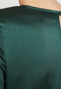 Mörkgrön, ribbad tyg med en slät textur. Har en nyckelhål detaljer vid bakre halsringningen och en lätt lyster i materialet.