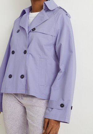 Persoon draagt een cropped lavendel trenchcoat met zwarte knopen over een wit shirt en lichtgrijze broek, staand tegen een witte achtergrond.