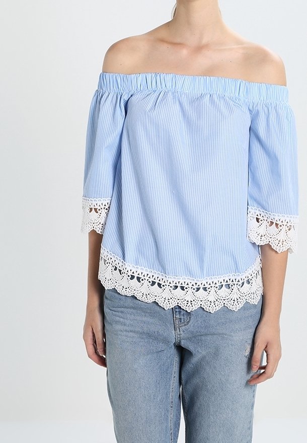 Femme portant un chemisier à rayures bleu clair, épaules dénudées, avec un galon en dentelle blanche sur les manches et l'ourlet, associé à un jean bleu clair.