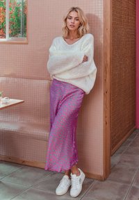 Young woman in white fuzzy sweater, purple polka dot skirt, and white sneakers leaning against a pink tiled wall indoors.