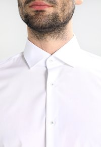 Biała koszula z gładkiej tkaniny. Posiada klasyczny kołnierzyk, zapięcie na guziki oraz subtelny połysk. Brak widocznych wzorów lub tekstur.