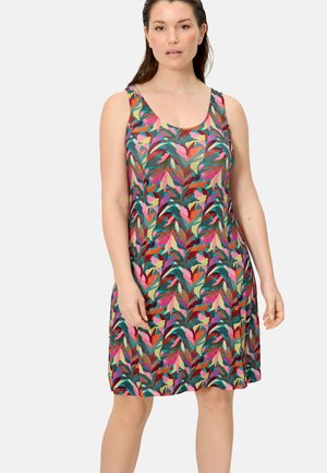 Mujer con vestido sin mangas hasta la rodilla con un patrón colorido de hojas tropicales, de pie contra un fondo claro y liso.