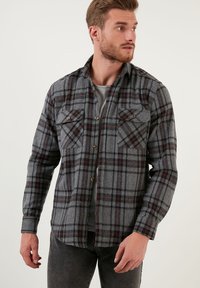 Camicia in flanella a quadri grigia con accenti rossi e neri. Presenta due tasche sul petto, chiusura con bottoni e maniche lunghe. Tessuto testurizzato.