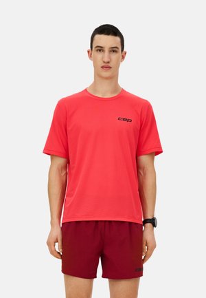 Man wearing a red short-sleeve sports shirt and dark red shorts, standing against a plain white background.