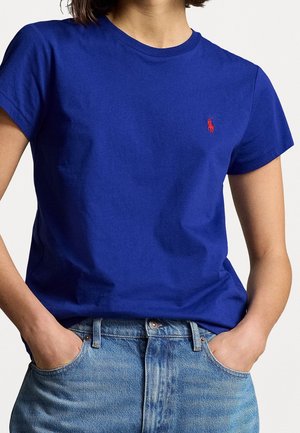 Person iført en kongeblå t-shirt med korte ærmer og et lille rødt broderet logo, kombineret med lyseblå denimjeans, hænderne i lommerne.