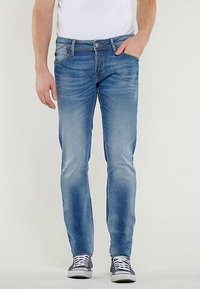 Lichtblauwe denim jeans met een slanke pasvorm, voorzien van voorzakken, subtiele vervaging en gestikte details. Gecombineerd met marineblauwe sneakers.