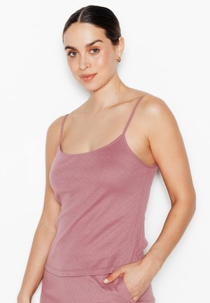 Femme aux cheveux foncés plaqués en arrière, portant un haut sans manches mauve et un bas assorti, posant avec une main dans la poche sur fond blanc.