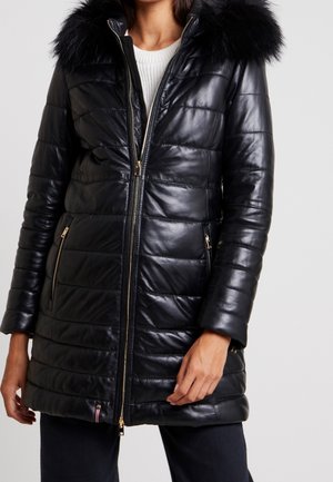 Vrouw draagt een zwart, doorgestikt leren jas met bontkraag, ritssluiting aan de voorkant, ritssluitingen op de zakken en een witte trui eronder.