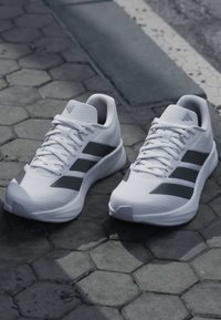 Paire de chaussures de sport blanches avec des rayures et des lacets noirs, posées sur un pavé en pierre hexagonal près du bord de la route.