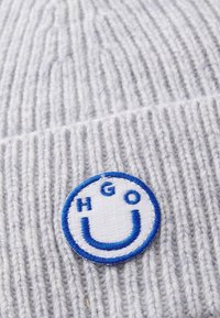 Grijze gebreide beanie met geribbelde structuur, met een ronde blauwe en witte geborduurde patch met het label "HGO" en een smiley-gezicht ontwerp.