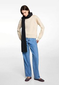Beige Pullover-Sweatshirt, dunkelgrauer Schal, hellblaue weite Jeans und schwarze gemusterte Slipper, getragen von einem Model.